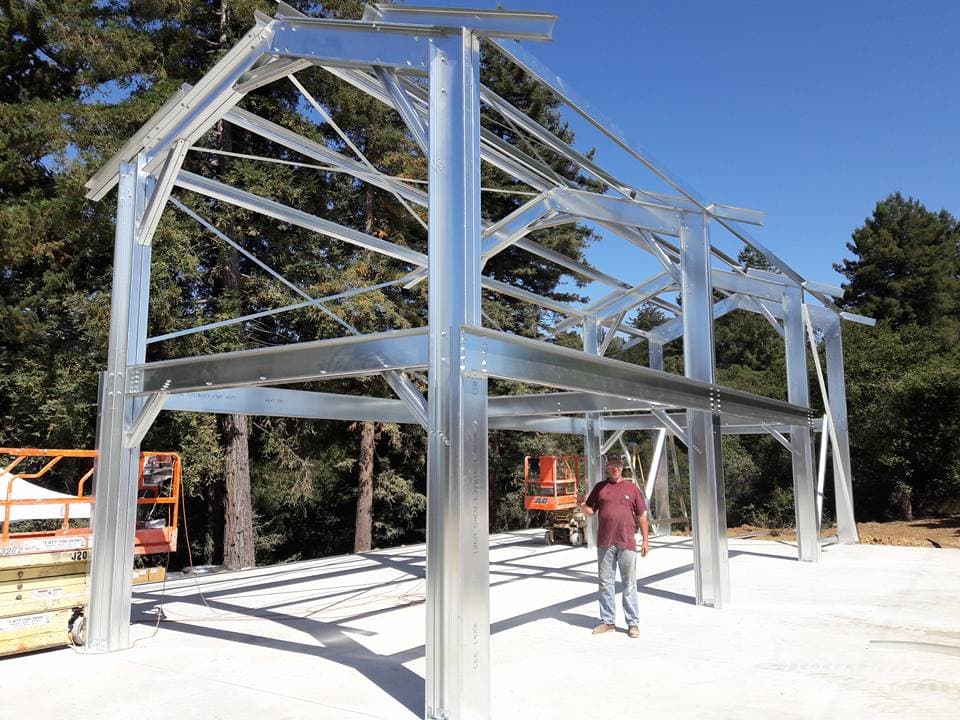 Man stands inside a large steel building frame under construction on a concrete foundation.