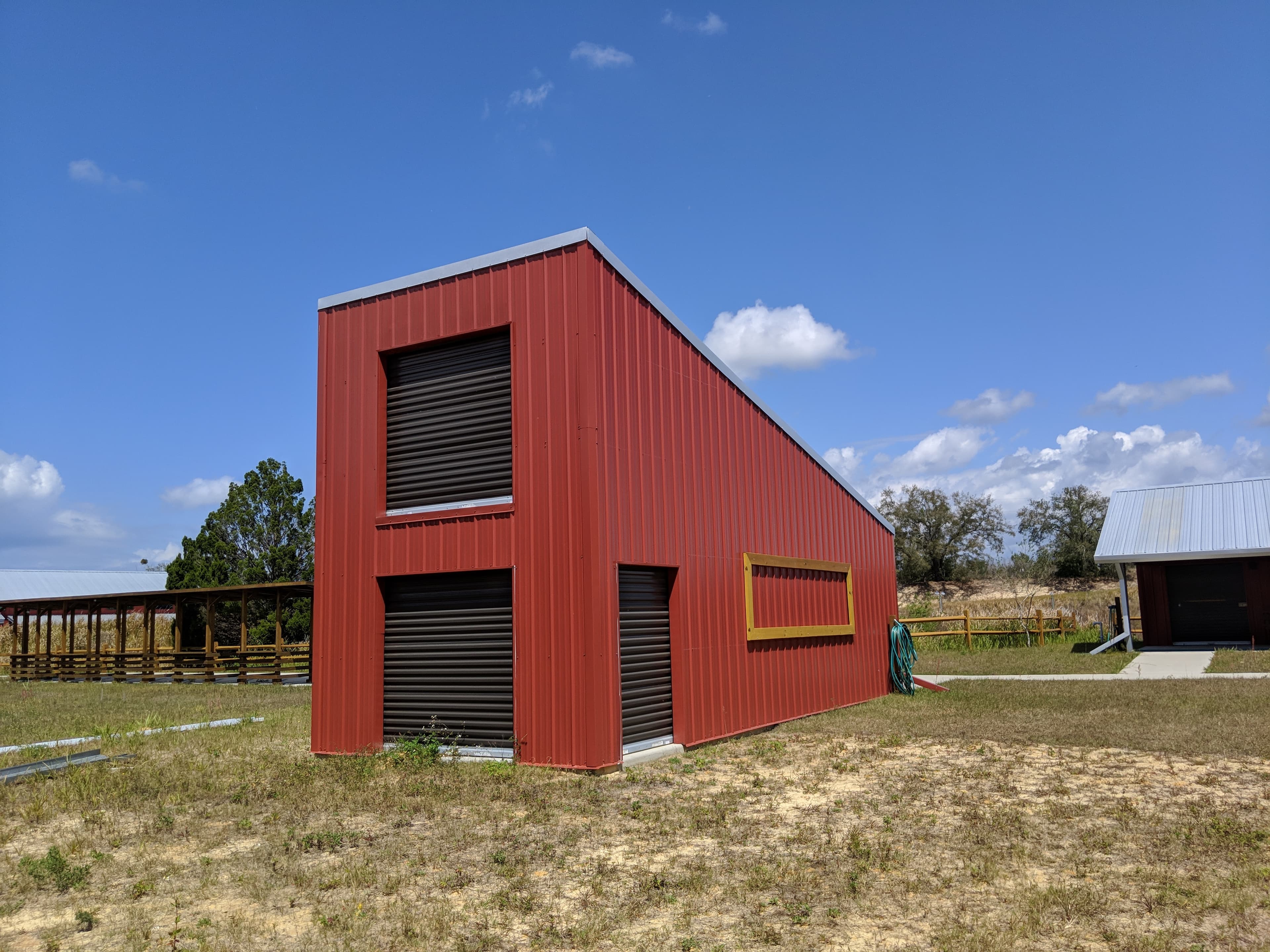 Red corrugated metal building with sloped roof and roll-up doors in a dry grassy field.