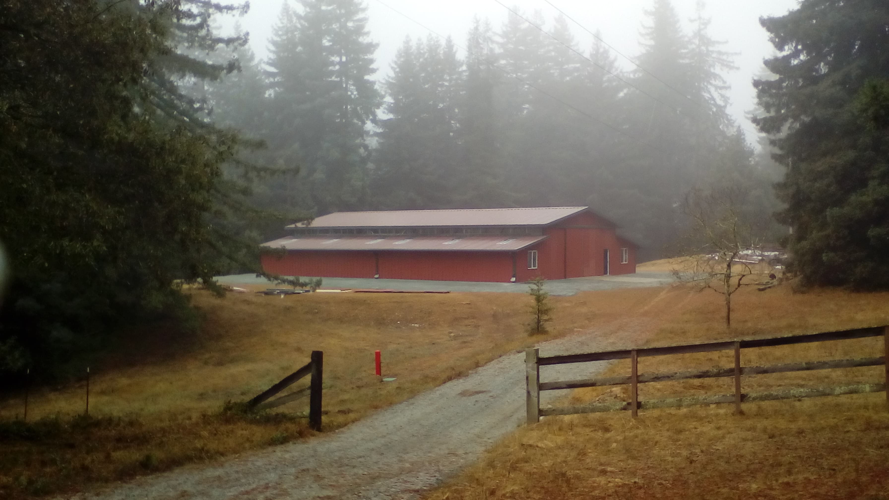 Large red barn in a foggy forest clearing with a gravel path and wooden fence.