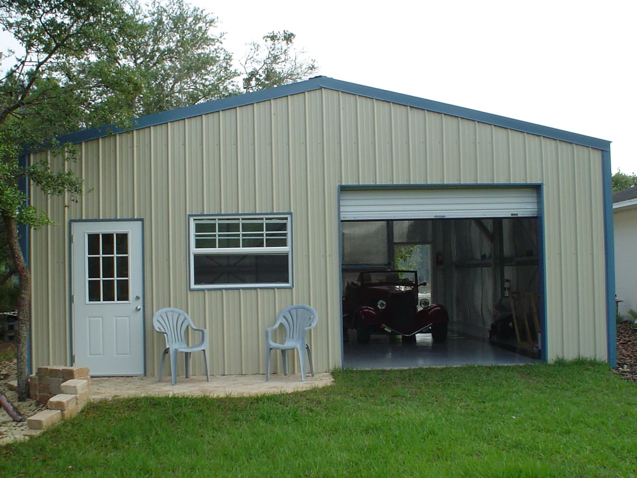 Tan metal garage with white door, window, and open bay showing a vintage red car.