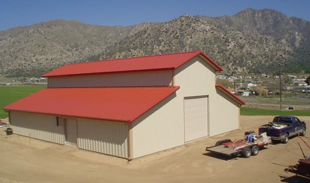 Large tan metal barn with a red roof and roll-up doors against rugged mountains.